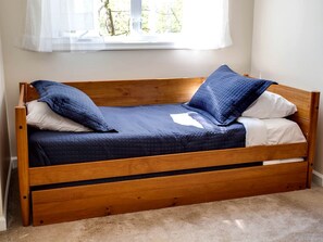 A wooden bed with a quilted blue mattress, white pillows, and a window with sheer curtains.
