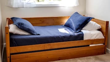A wooden bed with a quilted blue mattress, white pillows, and a window with sheer curtains.