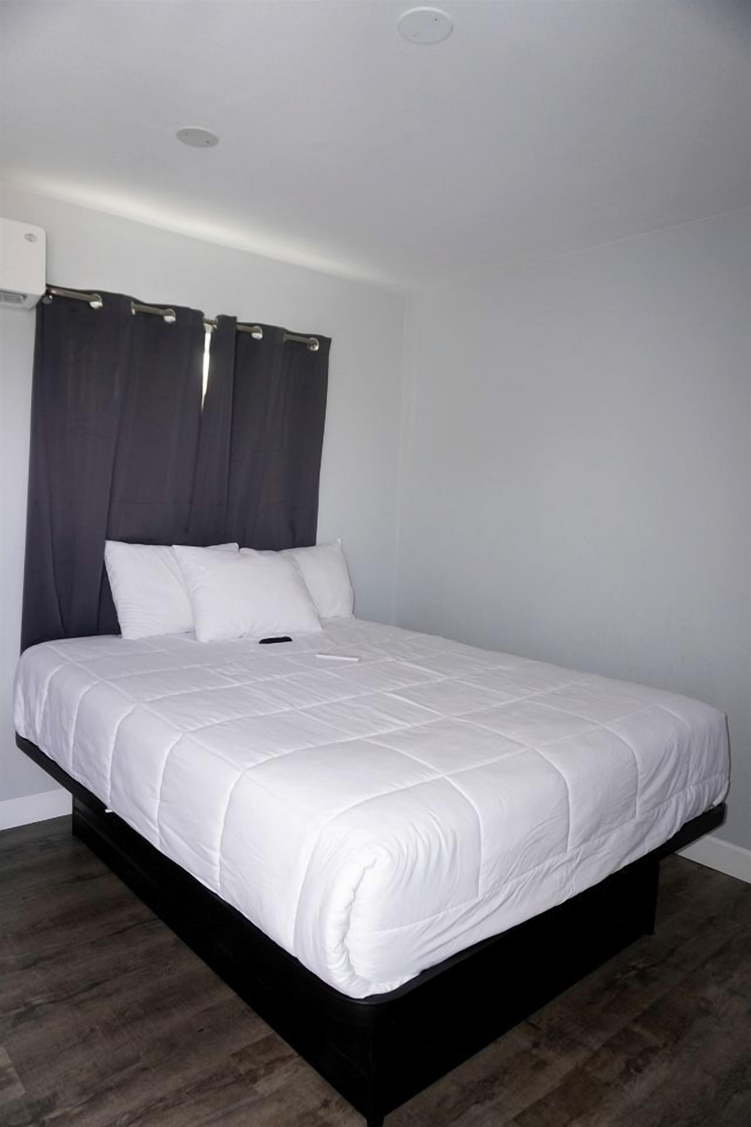 Un lit simple avec un matelas blanc et une tête de lit sombre, une petite table de chevet et un climatiseur fixé au mur.