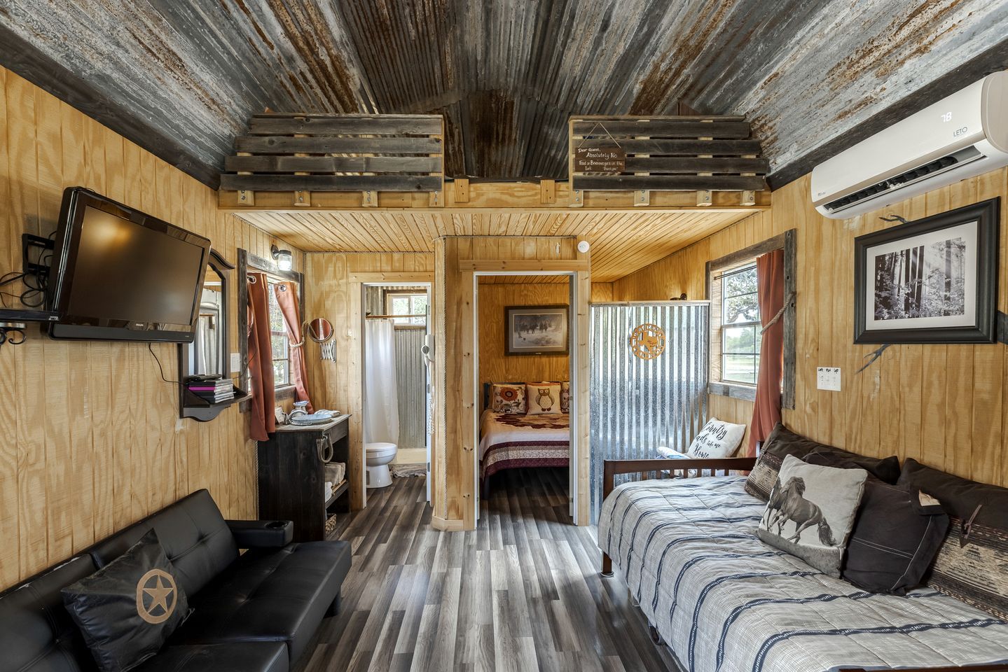 A rustic cabin interior with a wooden ceiling, a television, a bed with patterned bedding, a wooden bench, and a bathroom visible through an open door.