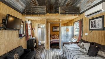 A rustic cabin interior with a wooden ceiling, a television, a bed with patterned bedding, a wooden bench, and a bathroom visible through an open door.