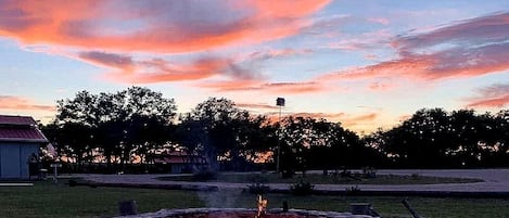 A stone fire pit with a fire burning, surrounded by a gravel area, under a sunset sky.