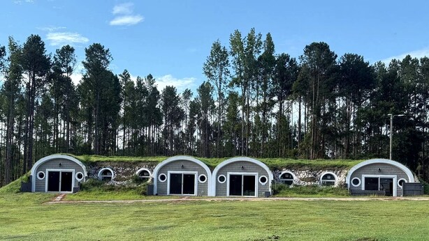 A row of four semi-circular buildings with large doors, set in a grassy field with a forest in the background. - Unique Earth Home Stay with Pet Goats and Fishing Pond near Lake Gaston NC (Littleton)