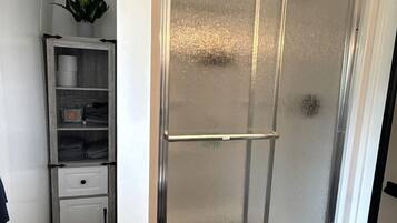 A bathroom with a glass shower enclosure, a white cabinet with shelves, and a small plant.