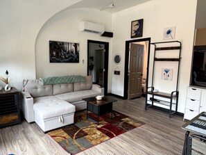 Ein Wohnzimmer mit Sofa, Couchtisch und Teppich. Eine Tür, ein Kleiderständer und ein kleiner Tisch mit einer Vase sind ebenfalls zu sehen.