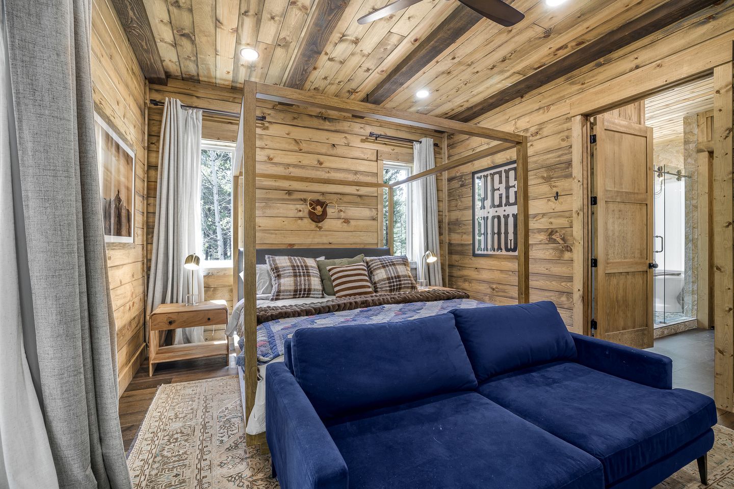 A wooden cabin interior with a bed, a blue sofa, a wooden bench, and a mounted deer head.