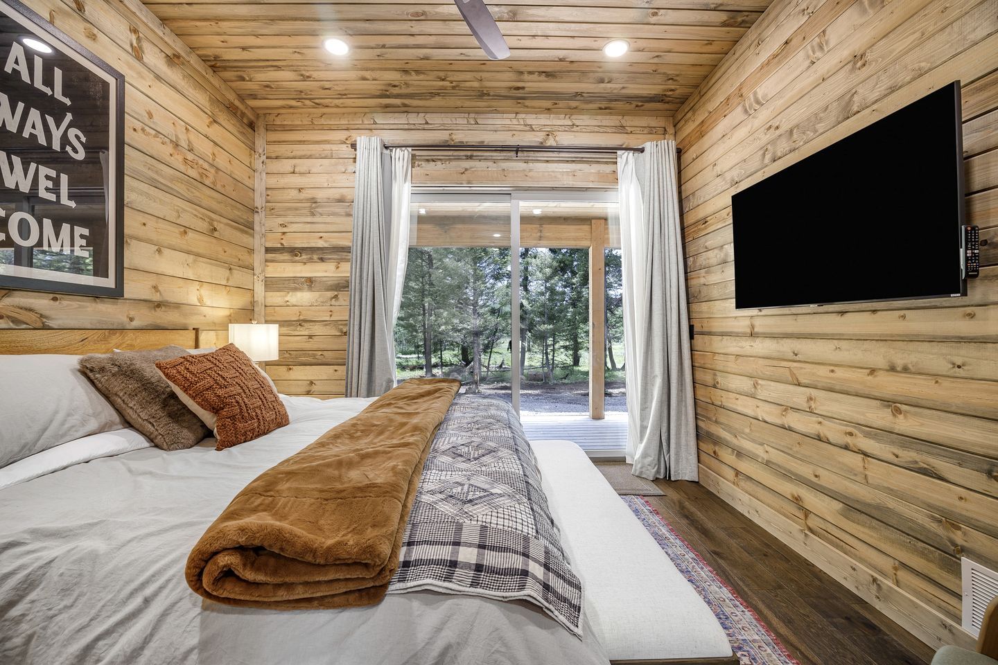 A wooden cabin bedroom with a bed, a television, and a large window.