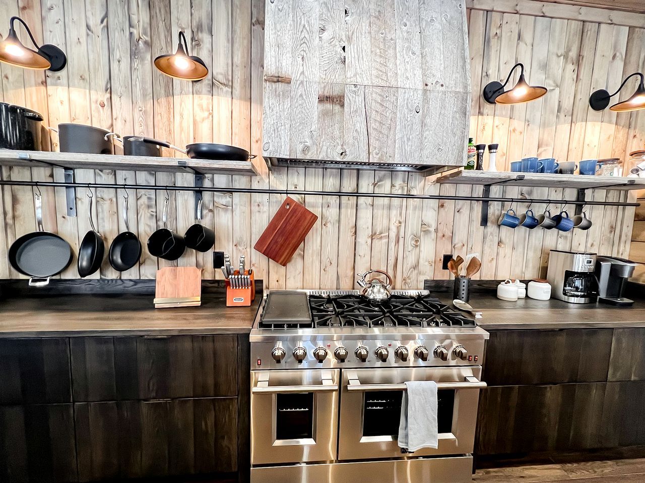 A kitchen with a stove, oven, hanging pots, and a wooden wall.