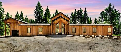 A wooden cabin with a large front door, a covered porch, and a dirt driveway.