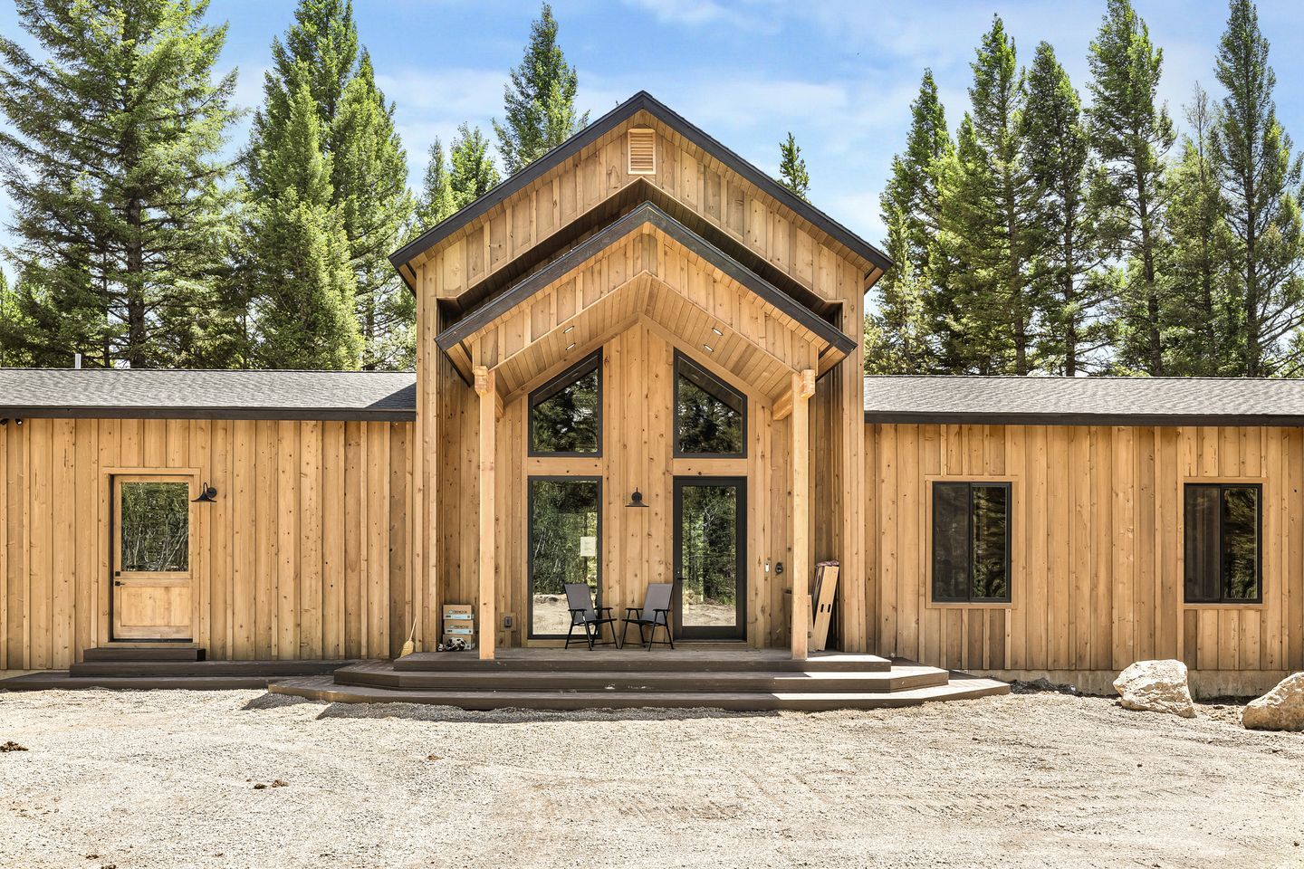 A wooden cabin with a central entrance, large windows, and a gravel driveway.