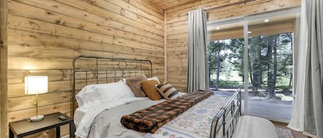 A wooden cabin bedroom with a bed, bedside table, lamp, and a view of the outdoors.