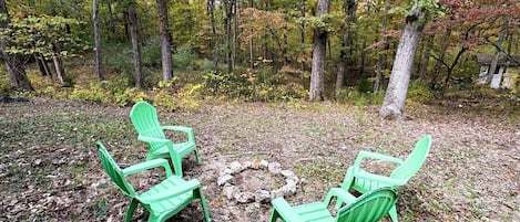 A forested area with three green Adirondack chairs arranged around a stone fire pit.