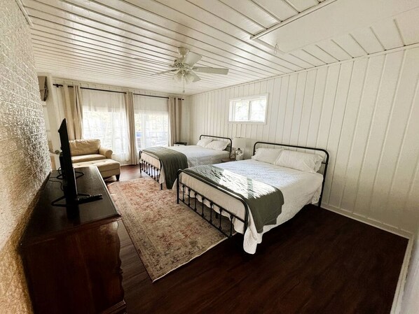 Un dormitorio con dos camas, un ventilador de techo, un televisor sobre una cómoda de madera y una ventana con cortinas.