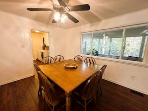 A dining room with a wooden table, six chairs, a ceiling fan, and a window with a view of greenery.
