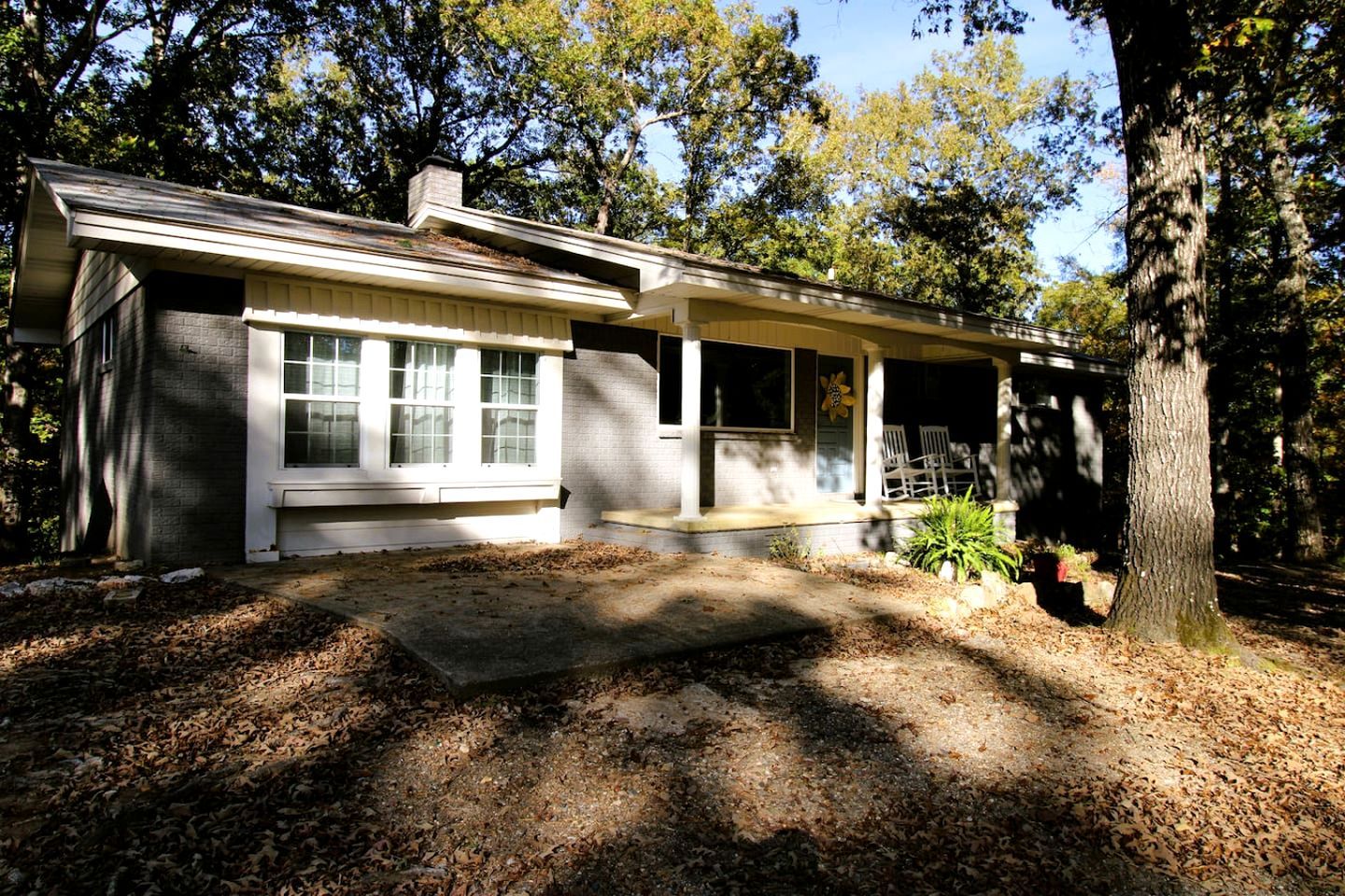 A single-story house with a porch, a window with blinds, and a tree in the foreground.