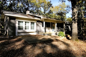 A single-story house with a porch, a window with blinds, and a tree in the foreground.