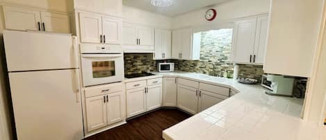 Una cocina con alacenas blancas, refrigerador, horno, microondas y salpicadero de azulejos.