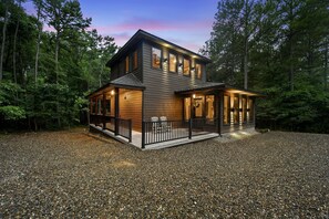 A two-story cabin with a covered porch, a gravel driveway, and surrounded by trees. - Woodland Romance (Broken Bow)