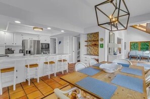 A modern kitchen with white cabinetry, a central island, and a dining area with a wooden table and blue placemats. - Riverfront Retreat: Hot Tub, Fire Pit, Private Dock & Screened Porch (Lancaster)