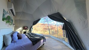 A tent-style room with a bed, a small table, and a window with curtains. - Luxury Geodome with Sauna, Hot Tub & Mountain Views near Pisgah National Forest, North Carolina (Balsam Grove)