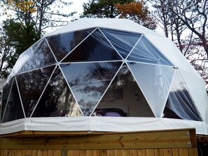 Uma estrutura em forma de cúpula geodésica com teto transparente, base de madeira e espaço interior visível.