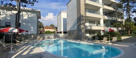 A modern building with a swimming pool, lounge chairs, and umbrellas.