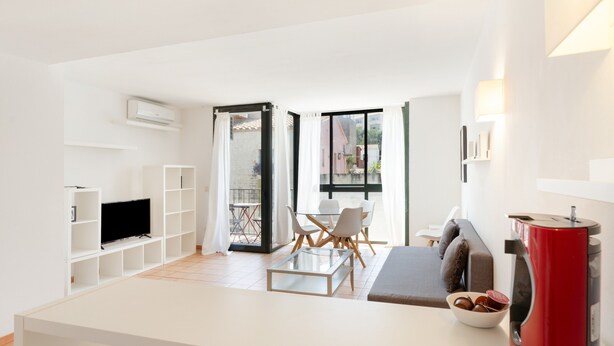 Ein modernes Wohnzimmer mit Fernseher, einem Essbereich mit Tisch und Stühlen, einer Couch, einer roten Kaffeemaschine und einem Balkon mit Blick.