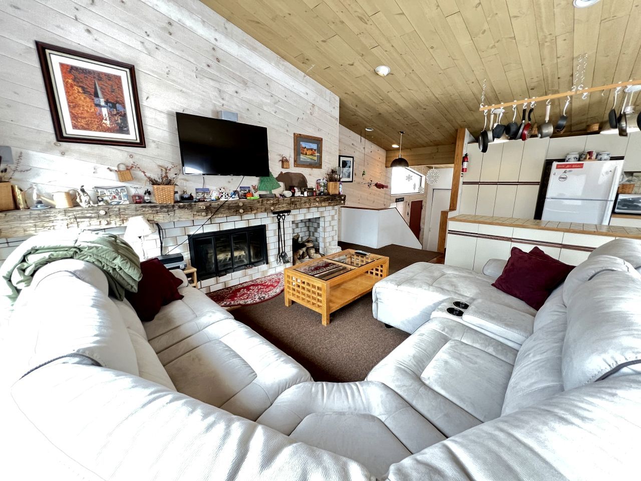 A cozy living room with a fireplace, a large flat-screen TV, and a kitchen area with hanging pots.