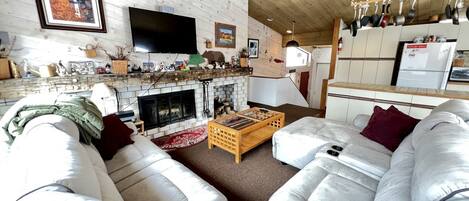 A cozy living room with a fireplace, a large flat-screen TV, and a kitchen area with hanging pots.