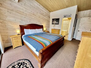 Un dormitorio con paneles de madera, una cama, mesitas de noche, un tocador y una puerta que da a otra habitación.