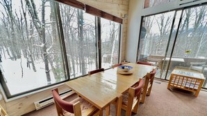 Une table à manger en bois, avec des chaises, de grandes fenêtres donnant sur la forêt et un petit tabouret en bois.