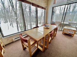 A wooden dining table with chairs, large windows with a view of trees, and a small wooden stool.