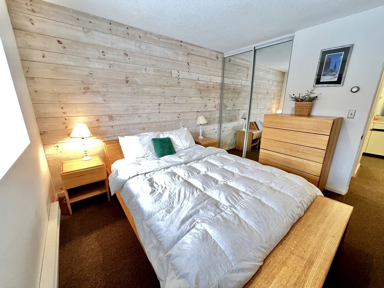 A bedroom with a wooden headboard, a bed with white bedding, a bedside table with a lamp, a dresser, and a mirror.
