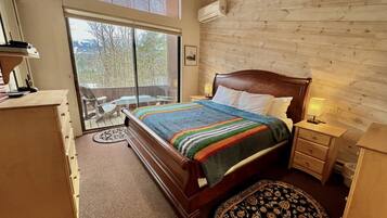 A bedroom with a wooden bed, a nightstand, a dresser, a window with blinds, and a view of trees outside.