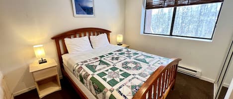 A bedroom with a wooden bed, a nightstand with a lamp, a window with blinds, and a framed picture on the wall.