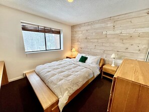 Un dormitorio con cabecera de madera, cama con ropa blanca, mesita de noche con lámpara y ventana con persianas.