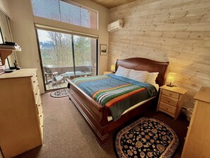 A bedroom with a wooden bed, a nightstand, a dresser, a window with blinds, and a view of trees outside.