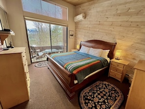 A bedroom with a wooden bed, a nightstand, a dresser, a window with blinds, and a view of trees outside.