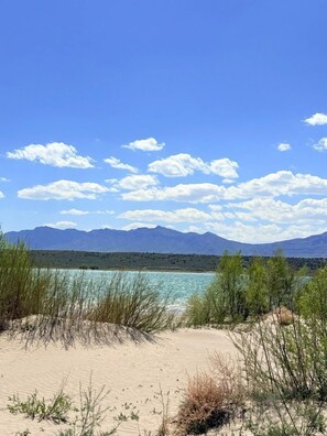 A beach with clear water, sand dunes, and mountains in the background.