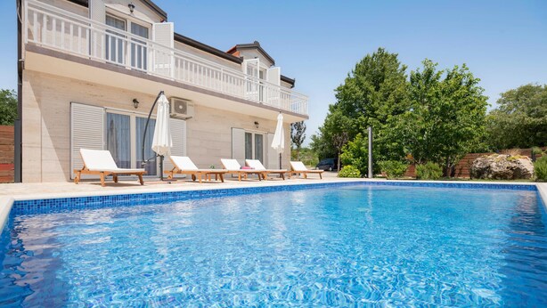 Une piscine avec des chaises longues et des parasols, un bâtiment doté d’un balcon et une végétation luxuriante.