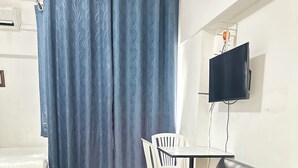 Une pièce comprenant un lit, un canapé en cuir noir, une table blanche avec des chaises, un téléviseur fixé au mur et un rideau bleu.