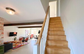 Eine Treppe mit Holzbodenstufen führt zu einer Zwischenebene, an einer Backsteinmauer vorbei, zu einem Wohnbereich mit Sofa, Sesseln und Fernseher.