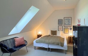 Ein Schlafzimmer mit Dachfenster, einem Bett mit Decke, Nachttischlampen, einem Sessel und Kunstwerken an den Wänden.
