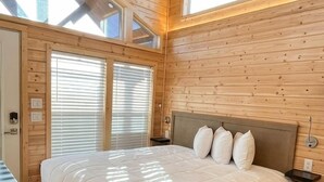 Et soveværelse i en træhytte med seng, ventilator og store vinduer.