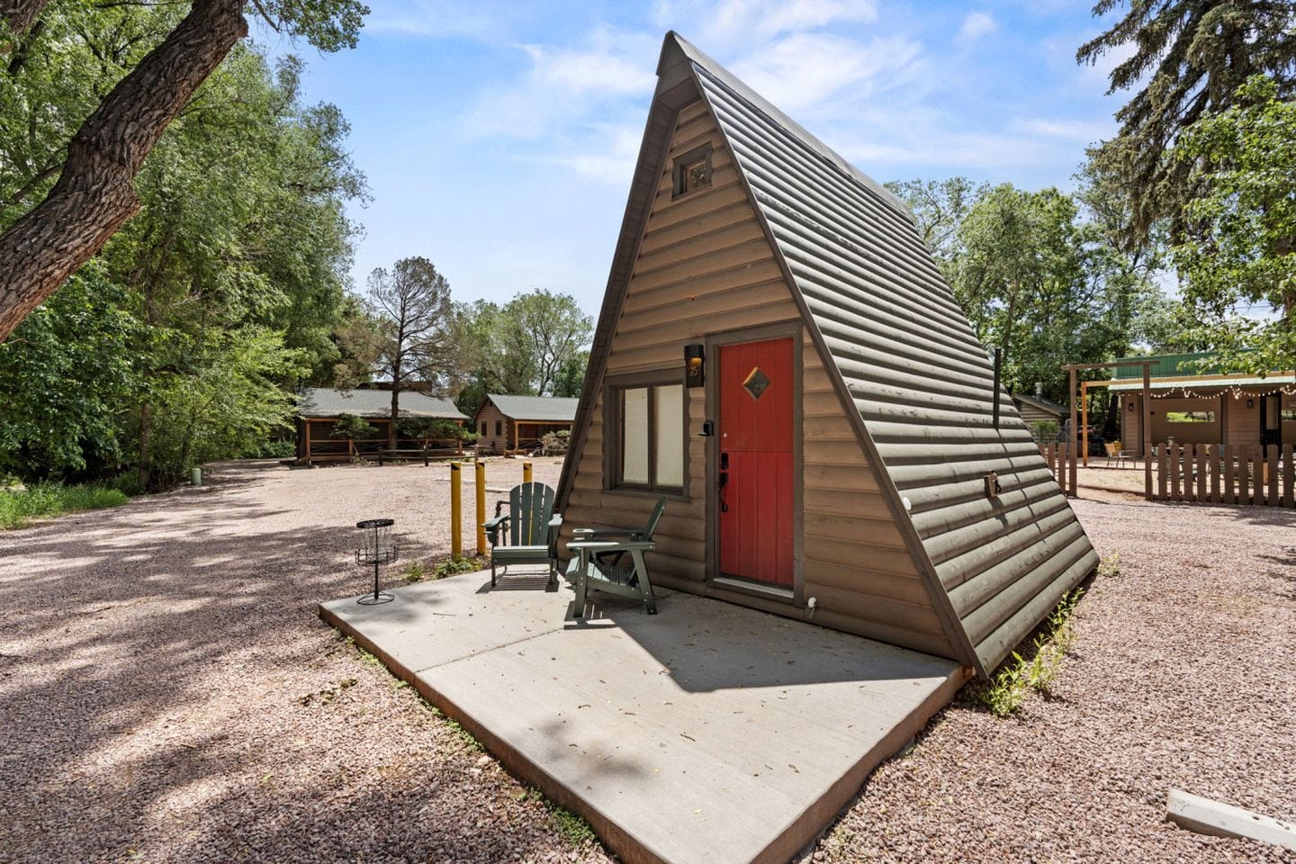 A triangular wooden cabin with a red door, a concrete patio, and a gravel ground.
