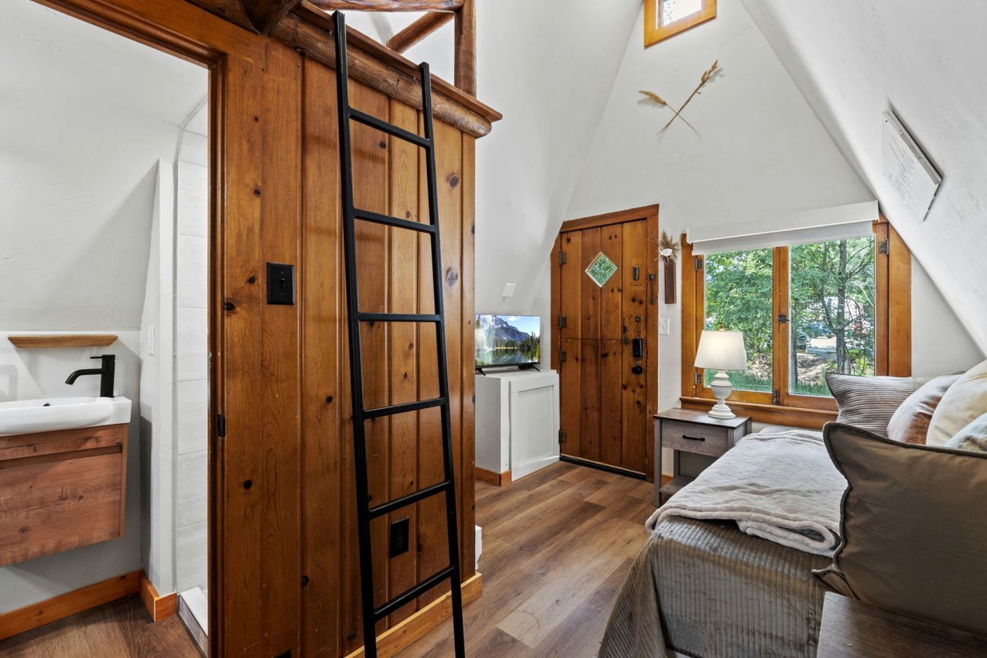 A loft-style bedroom with a wooden ladder, a bed, a bedside table with a lamp, and a view of the outdoors through a window.