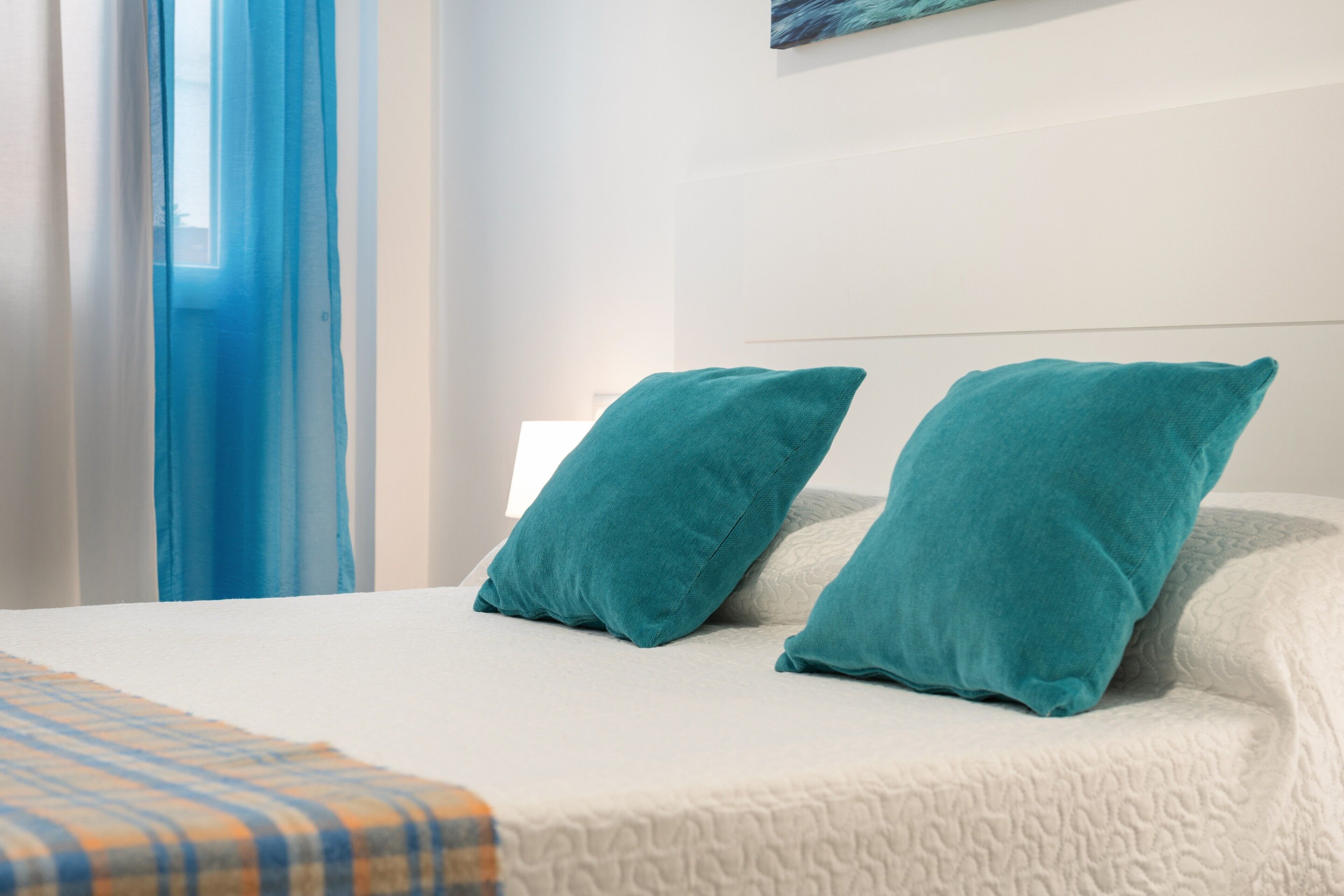 Un lit recouvert d’une couverture blanche à texture, agrémenté de deux coussins turquoise et d’une couverture à carreaux.