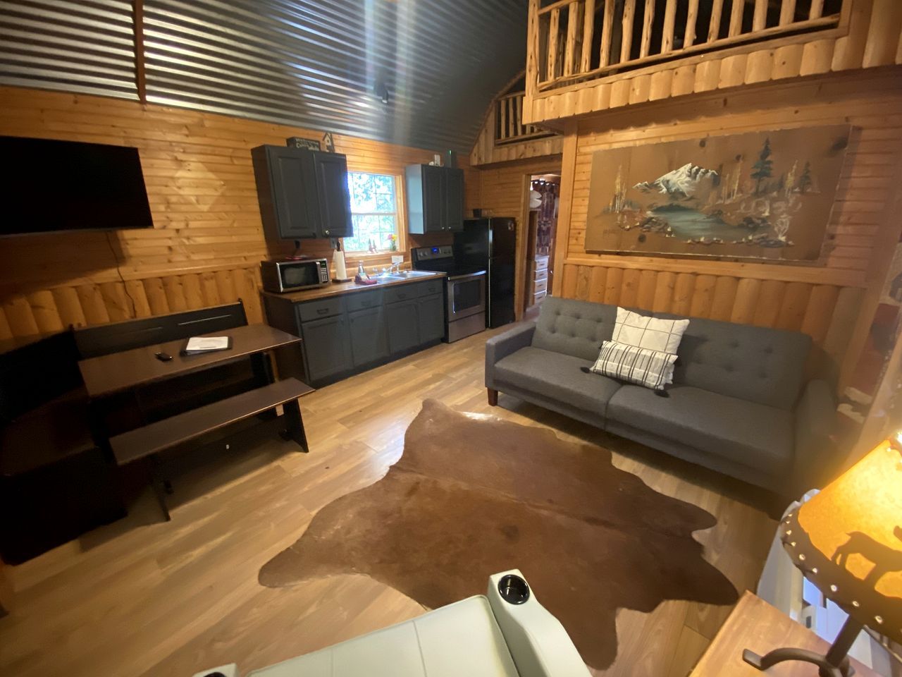 A cabin interior with a wooden floor, a sofa, a dining table, a kitchen area, and a mounted television.