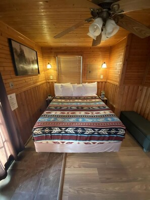 Dormitorio de cabaña de madera con cama, dos luces empotradas en la pared, ventilador de techo y ventana con persianas.