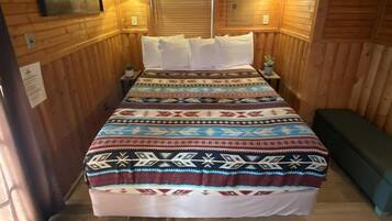 A wooden cabin bedroom with a bed, two wall-mounted lights, a ceiling fan, and a window with blinds.